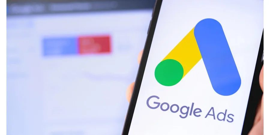 Phone held in a hand showing the Google Ads Logo with a blurred versions of the google ads interface in the background